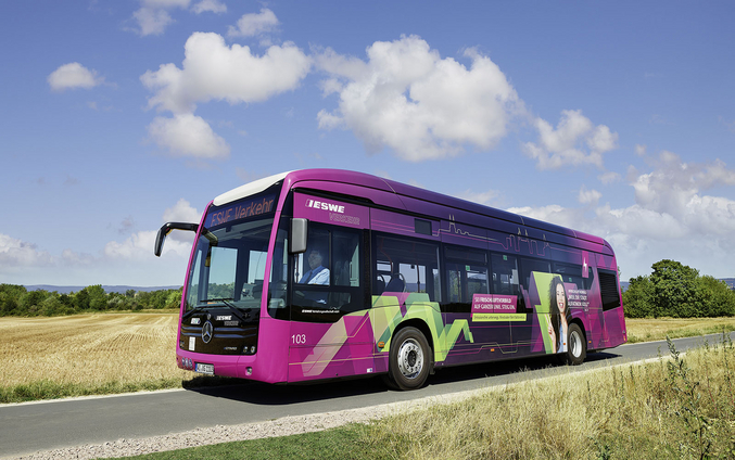 Un autobús eléctrico ESWE de color púrpura en un camino de campo contra un cielo azul.