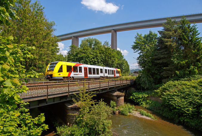 Un tren HLB blanco y amarillo cruza un puente que pasa por encima de un río en un bosque.