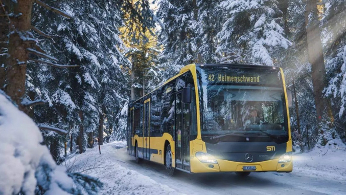 Ein gelber Bus der STI Bus AG fährt durch einen verschneiten Tannenwald.