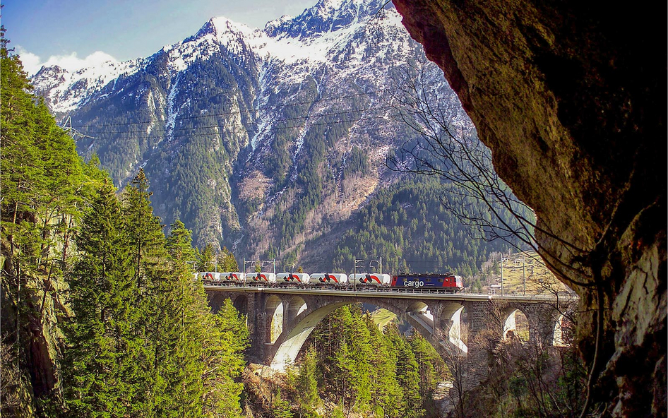 Un ferrocarril cruza un puente de izquierda a derecha ante un panorama montañoso.