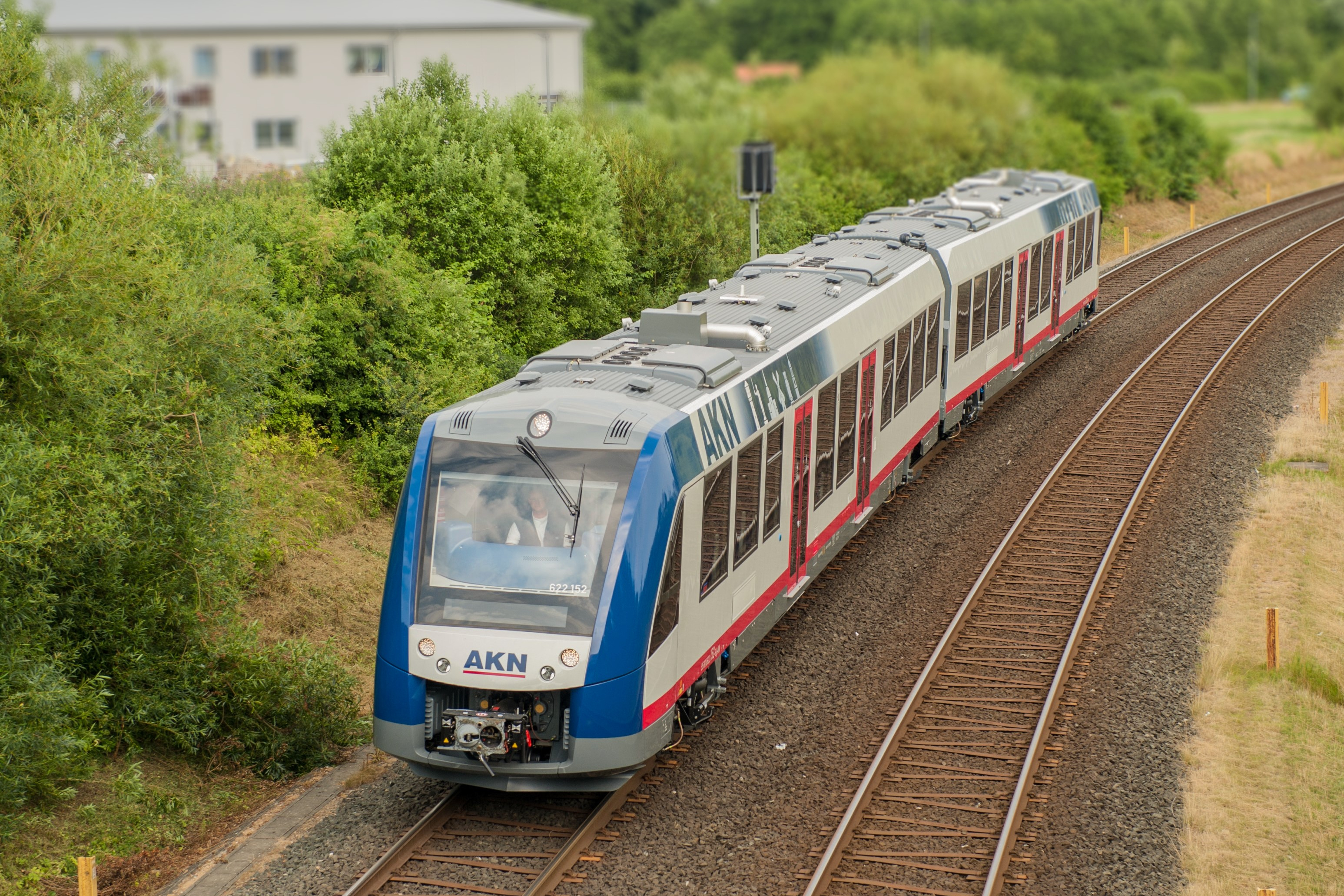 Ein blau-weiß-roter AKN Zug fährt nahe einer Ortschaft im Grünen.