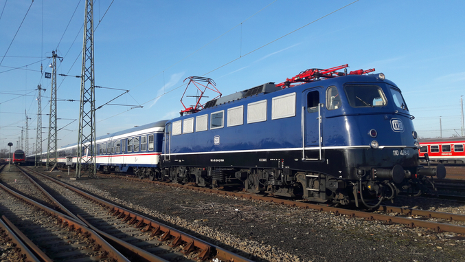 Un tren TRI azul circula de izquierda a derecha sobre raíles marrones.